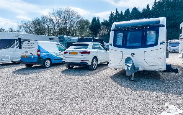 A white and blue RS Connect van parked next to a Coachman caravan in a sunny motorhome park, with other motorhomes visible in the background. An RS Connect engineer is installing a tracker inside the caravan.
